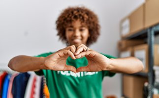 Young african american woman wearing volunteer uniform doing heart symbol with hands at charity centre