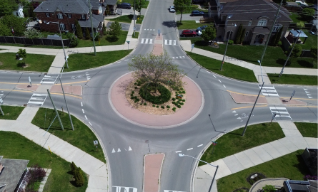 A roundabout in a residential area with houses nearby and sidewalks on either side.
