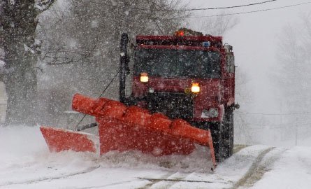 Snow plow pushing snow off a rural road on a snowy overcast day.